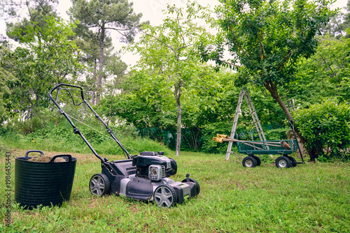 Gasoline lawn mower parked on grass next to a basket for waste, a garden cart and a step ladder used for maintenance tasks in a large rural garden.