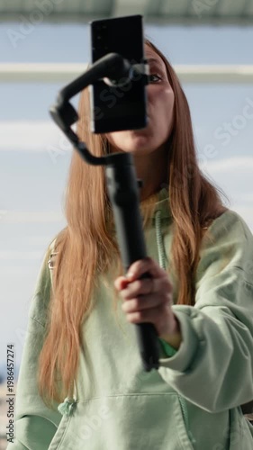 Vertical Video Girl content creator using phone on tripod to record vlog in airport lounge, capturing travel journey before boarding. Busy terminal illustrates social media presence and mobile filming