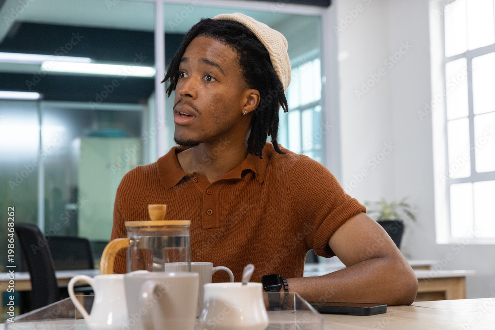 Fototapeta premium African American man 20s wearing knit hat rust polo sitting in office pouring from glass teapot