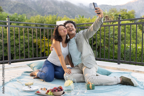 Romantic couple sitting on blue picnic blanket on rooftop terrace, taking selfie with smartphone