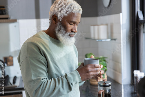 Senior African American man standing in kitchen wearing sage sweater holding mug near French press