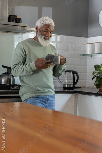 Senior African American man leaning on black counter in kitchen, holding smartphone and gray mug