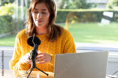 Woman recording at wooden table beside window, wearing yellow sweater and white headphones with mic