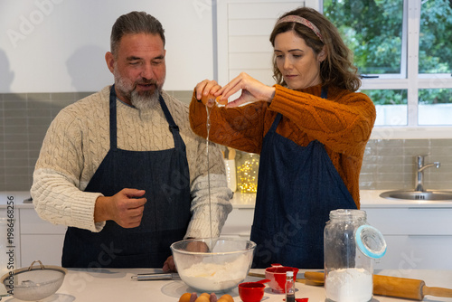 Couple in aprons preparing baking mix in home kitchen, cracking egg into glass bowl