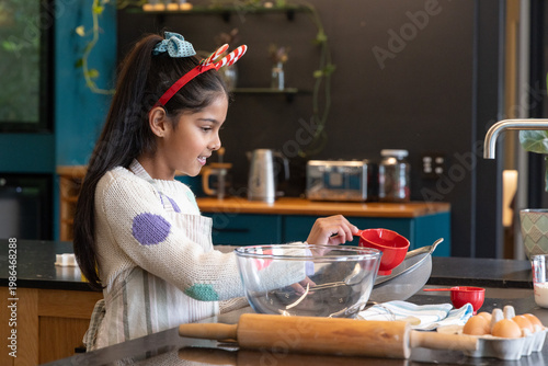 Asian female child in polka-dot sweater and striped apron, measuring with red cup over glass bowl