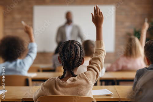 Young girl raising her hand in a diverse classroom with a teacher in the background, concept of education, active participation, and inclusive learning environments.
