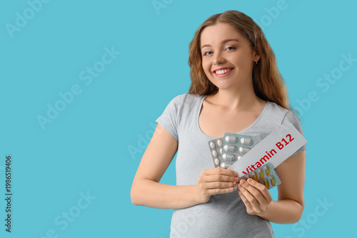 Young pregnant woman holding paper with text VITAMIN B12 and pills on blue ba...