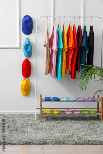 Cuadro en lienzo Rack with colorful t-shirts, caps and flip-flops near light wall