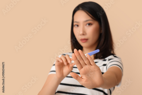 Beautiful young Asian woman with pregnancy test showing stop sign on beige ba...