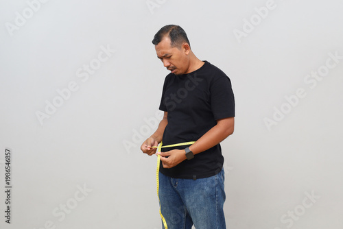 Obraz na płótnie Standing side view of a man measuring his waist or measuring his stomach circumference with a measuring tape, disappointed expression, concept of failed diet or obesity