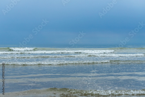 Calm ocean waves rolling toward the shore under a pale blue sky in a tranquil...