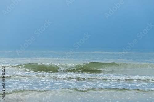Calm ocean waves roll toward the shore beneath a pale blue sky, forming a tra...