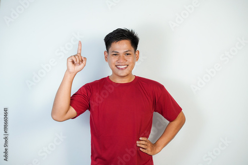 Smiling young Asian man wearing a red shirt pointing upward against a plain white background.