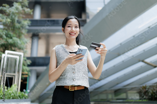 A woman smiles outdoors while holding a smartphone and a credit card, appearing to make an online transaction in a modern urban setting.