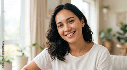 Free Download A smiling woman with wavy dark hair sits indoors, looking directly at the camera. She wears a simple white shirt and has a warm, genuine expression. - GFXLoad