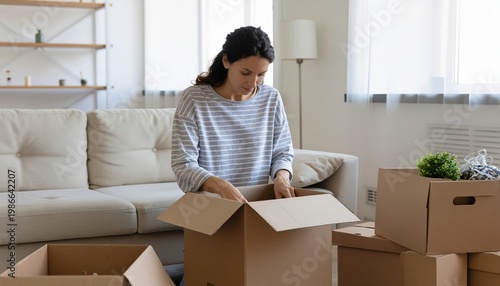 A woman carefully unpacks items from a cardboard box, with a couch and other boxes nearby. This suggests the woman may be moving, settling into a new home, or organizing her belongings.