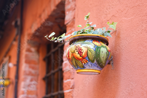 Traditional hand painted Italian ceramic flower pot with ivy on terracotta wall. Close-up shot features colorful Mediterranean pottery with pomegranate and cactus motifs and copy space. Authentic