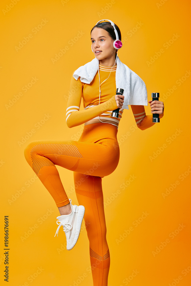 Naklejka premium Woman fitness model in orange athletic wear pumps movement with dumbbells, towel and headphones against a vivid orange studio backdrop.