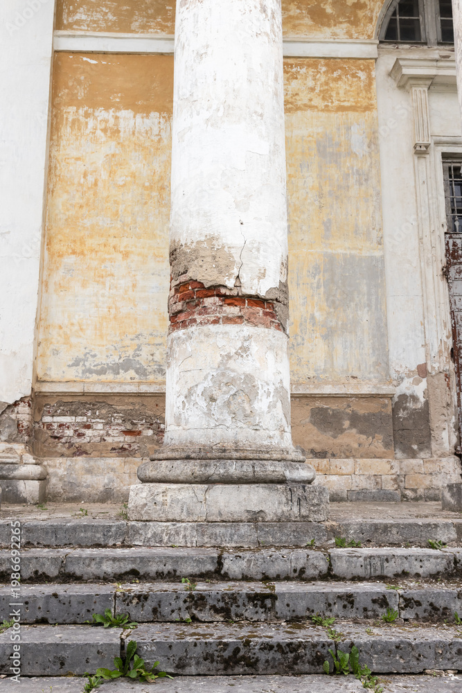 Fototapeta premium A weathered classical column stands on mossy stone steps in front of a historic building with peeling plaster, exposed brick and a crumbling facade, evoking age, decay and architectural heritage