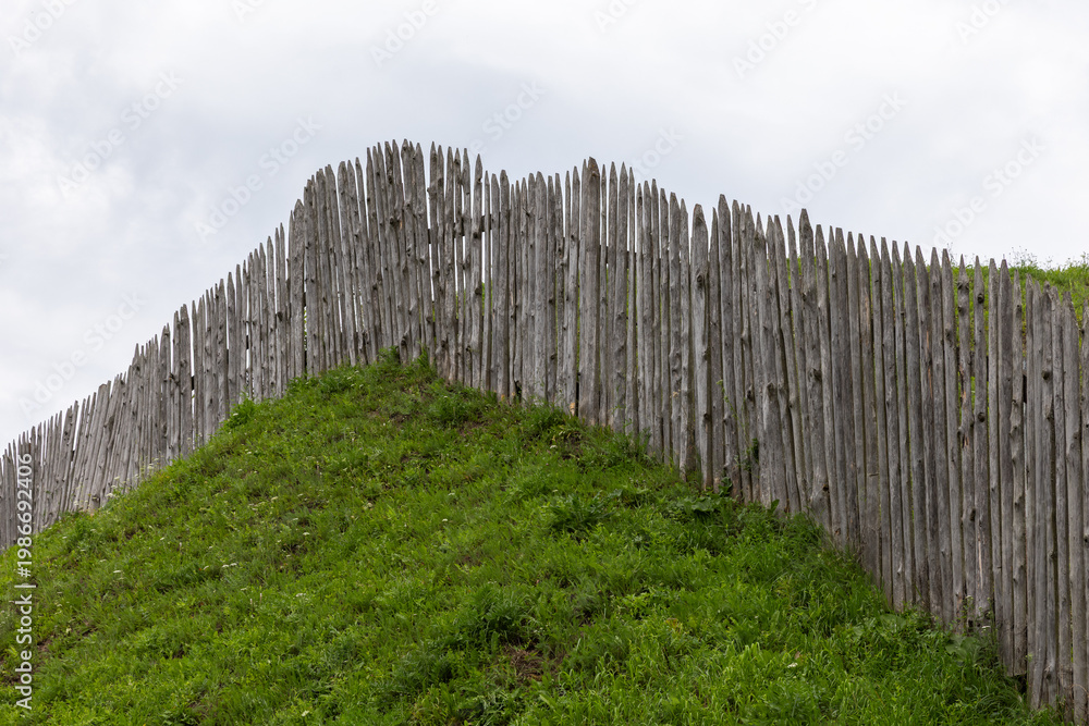 Fototapeta premium A weathered wooden palisade fence crowns a grass-covered earthwork hill beneath an overcast sky, suggesting rustic fortification, historic architecture and rural landscape textures with green grass
