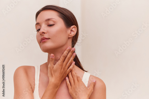 Skin care woman applying neck cream with both hands gently pressing her neck and décolleté, eyes closed in a serene relaxed expression. Clean beige background, copy space right. Ideal for neck cream.