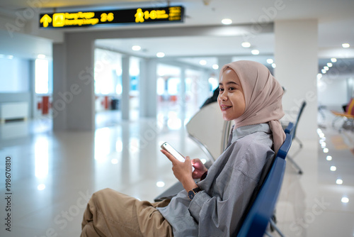 Muslim woman using smartphone while waiting in airport terminal seat