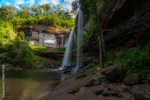 A waterfall cascading from a high cliff in the middle of the forest, with a sandy bottom and trees.