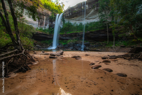 A waterfall cascading from a high cliff in the middle of the forest, with a sandy bottom and trees.