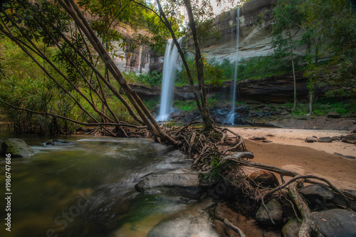 A waterfall cascading from a high cliff in the middle of the forest, with a sandy bottom and trees.