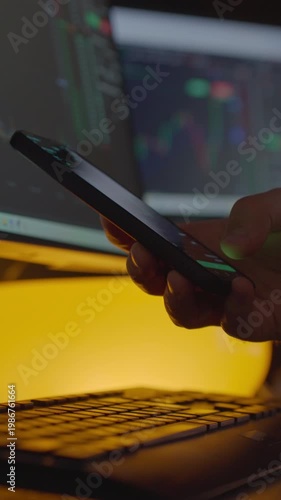 Trader using mobile app with financial chart while working near laptop in the night. Hand holding smartphone with stock market chart