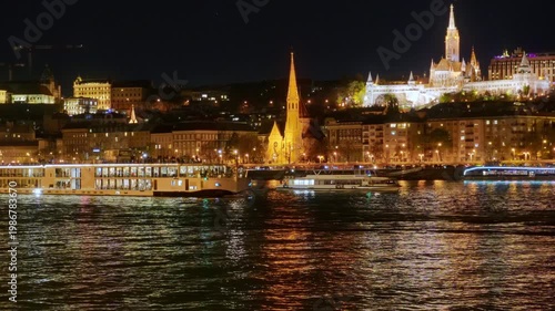 architecture with warm golden illumination reflecting in the river water at night, featuring passing cruise boats in Budapest city