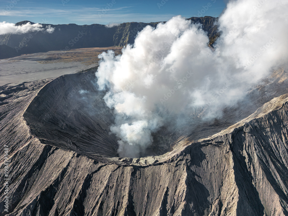 Obraz premium Aerial view of the Bromo volcano's caldera emitting a plume of white smoke, a stark contrast against the dark, rugged terrain, Mt. Bromo, East Java, Indonesia