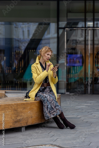 Adult female in yellow coat using touch screen of smartphone while sitting on urban wooden bench