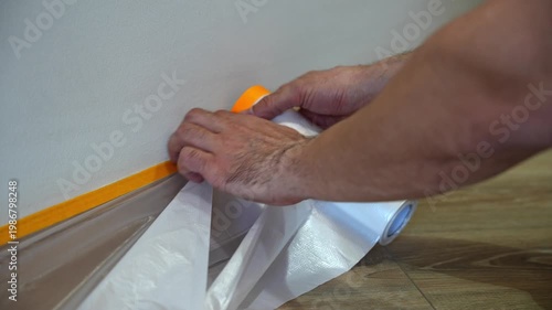 Closeup view of male hands applying protective film with adhesive tape along baseboard to protect floor during wall painting at home.