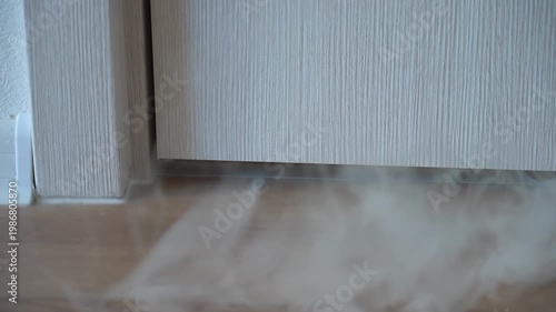 Smoke starts coming from under the door concept of fire smoke ignition. The living room has a close-up view of the entrance door, from under which thick white smoke begins to abruptly rise. High
