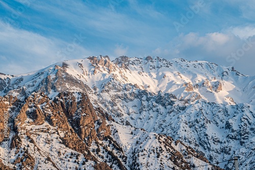 A stunning view of a snow-covered mountain range, showcasing rugged peaks and rocky terrain against a clear blue sky with wispy clouds.