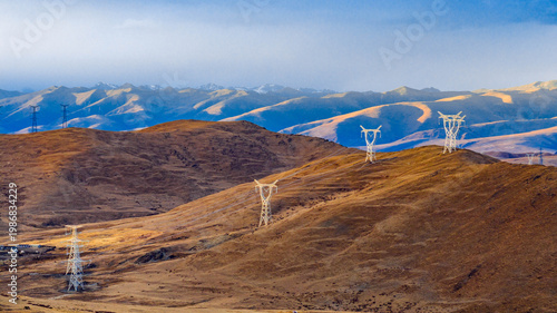 High-voltage power transmission towers and lines extending across barren mountains in western China. The scene illustrates electricity infrastructure in remote and arid regions.