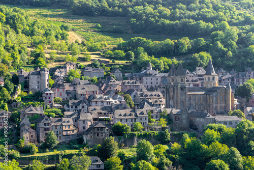 Medieval village of Conques perched on a green hillside at golden hour, panoramic view from a hill, soft evening light, France