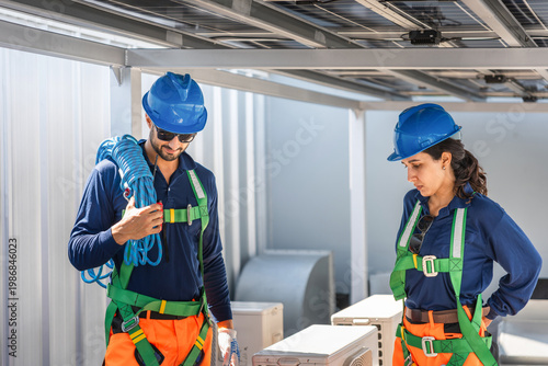 Engineers in safety harness and hardhats inspecting solar panel installation on rooftop, Sustainable energy technicians team installing solar power system on industrial building rooftop