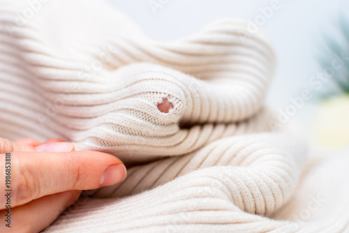 Clothing repair, craft sewing and textile recycle. Woman hands holding light beige knitted texture cloth with hole made by moth or ripped, close-up. Tailor holding damaged fabric with loose loops.