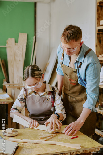Obraz na plátně Vertical shot of teenage girl woodworking with middle aged Caucasian male instructor, measuring timber in workshop