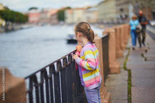Young girl holding a doll and looking at the Fontanka River from the embankment in Saint Petersburg, capturing childhood, travel and city life in Russia