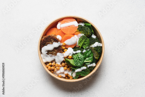 Healthy vegan lunch with broccoli, roasted chickpeas, spinach, carrot, lentil patty and yogurt dressing in round cardboard container on white background with copy space