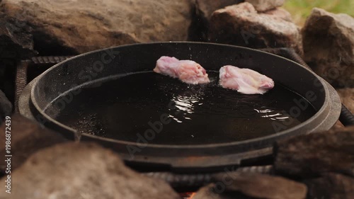 Chef placing raw chicken wings into a hot frying pan with oil, cooking meal on open fire outdoors among rocks.