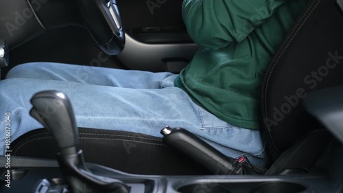 A man wearing a red plaid shirt is fastening his seatbelt in his private car. This is a safety precaution before driving.
