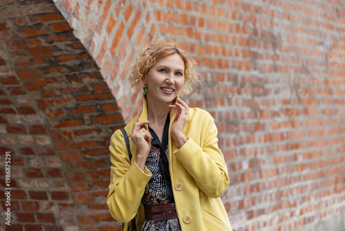 Close up of smiling mature woman in yellow coat touching collar by red brick background