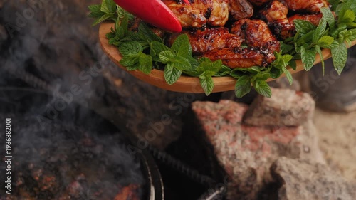 Roasted BBQ chicken wings on a wooden platter with fresh mint leaves, chef taking meat off the fire with tongs.