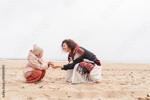 Tableau sur toile Woman showing seashells to a little girl while sitting on a sandy beach, educational moment, motherly care, seaside discovery