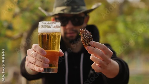 Man in a hat holding a glass of light beer and a fried chicken wing with sesame seeds in an autumn park.