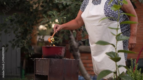 Woman in apron cooking vegetable stew in a pot on a grill, stirring ingredients with a wooden spoon over an open fire.